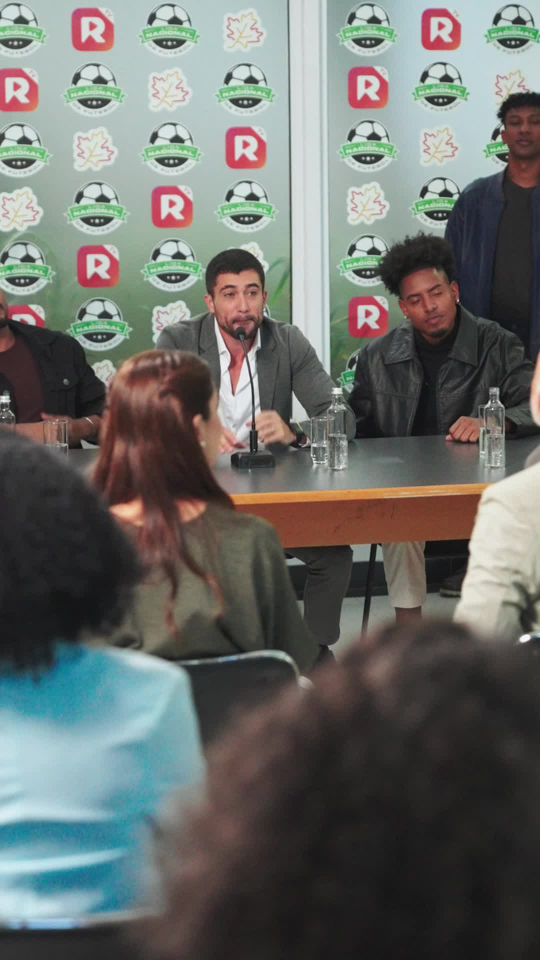 Coletiva de imprensa da Liga Nacional de Futebol: Lucas Moreti de blazer cinza fala ao microfone entre outros jogadores, com uma mulher de costas na primeira fila