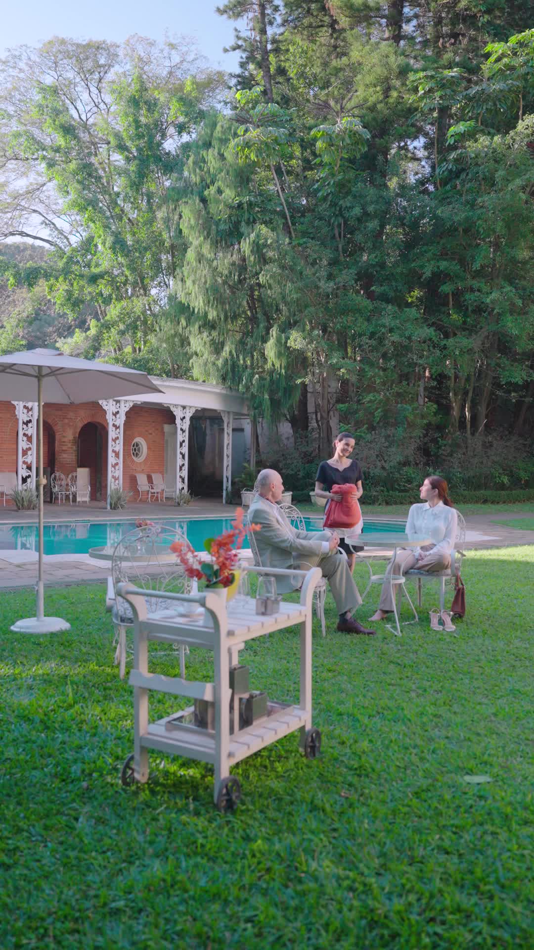 Raquel Falcão de blusa branca sentada a uma mesa à beira da piscina de uma mansão, conversando com um homem mais velho de terno claro