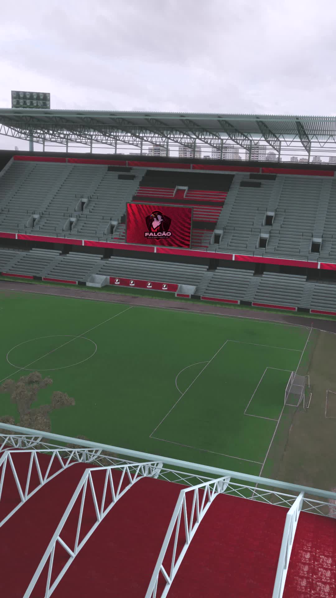 Estádio de futebol vazio, com arquibancadas cinza e vermelhas e um telão com o escudo do Falcão Sociedade Esportiva