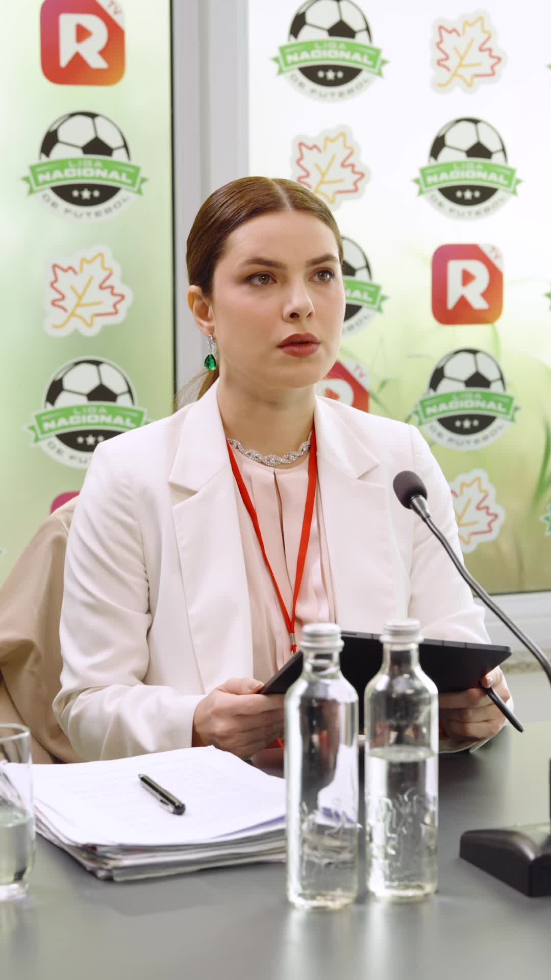 Raquel Falcão de blazer branco, brincos de esmeralda e crachá, sentada diante de um microfone com um tablet, em frente ao painel da Liga Nacional de Futebol