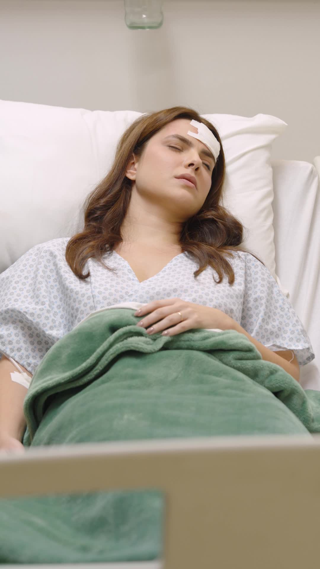 Raquel Falcão deitada numa cama de hospital, de camisola hospitalar, com um curativo na testa e os olhos fechados