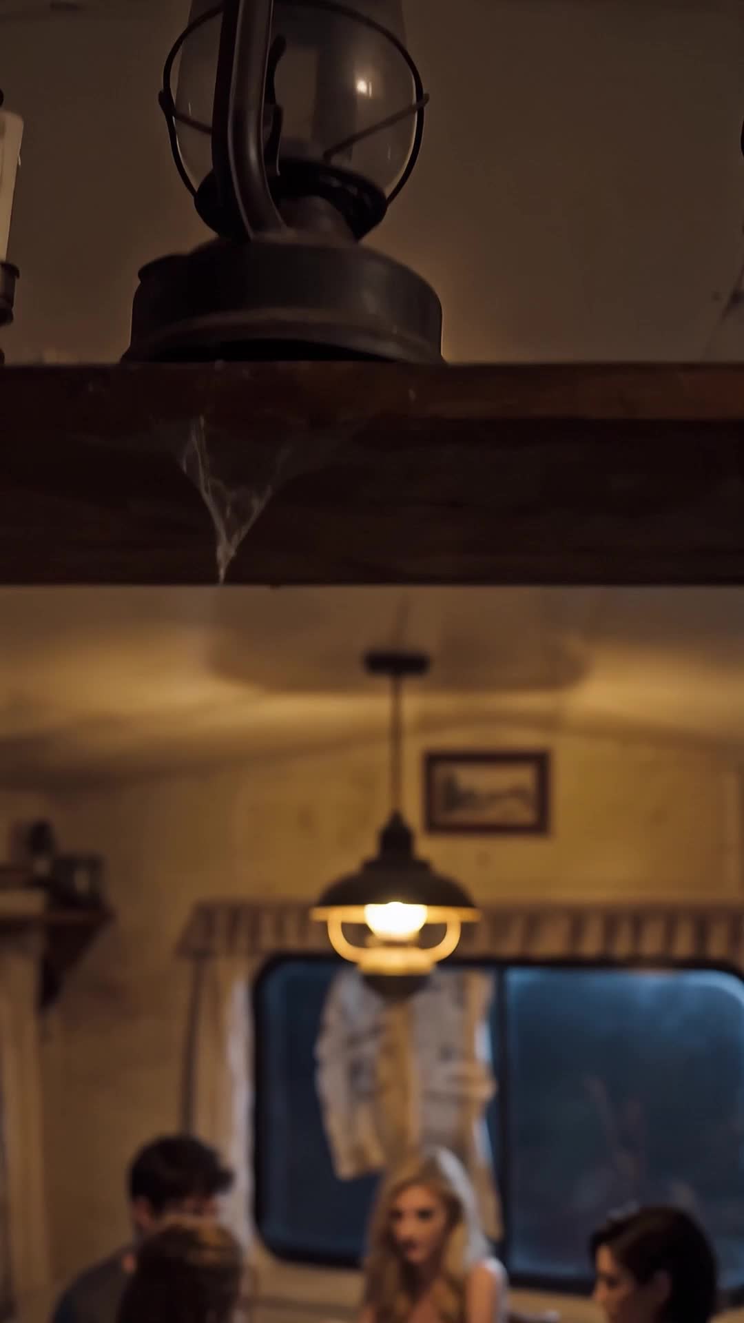 Um lampião de metal com cúpula de vidro sobre uma prateleira de madeira com teia de aranha e, desfocados abaixo, uma luminária acesa pendurada, uma janela e três pessoas à mesa, entre elas uma loira de blusa clara e um homem de cabelo escuro