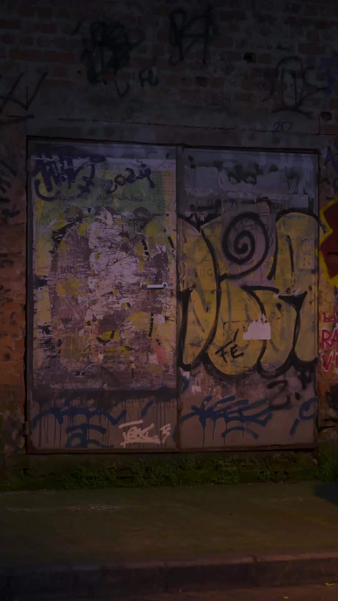Porta dupla de metal coberta de pichações e cartazes rasgados num muro de tijolos, à noite, com mato no chão e a luz amarela de um poste