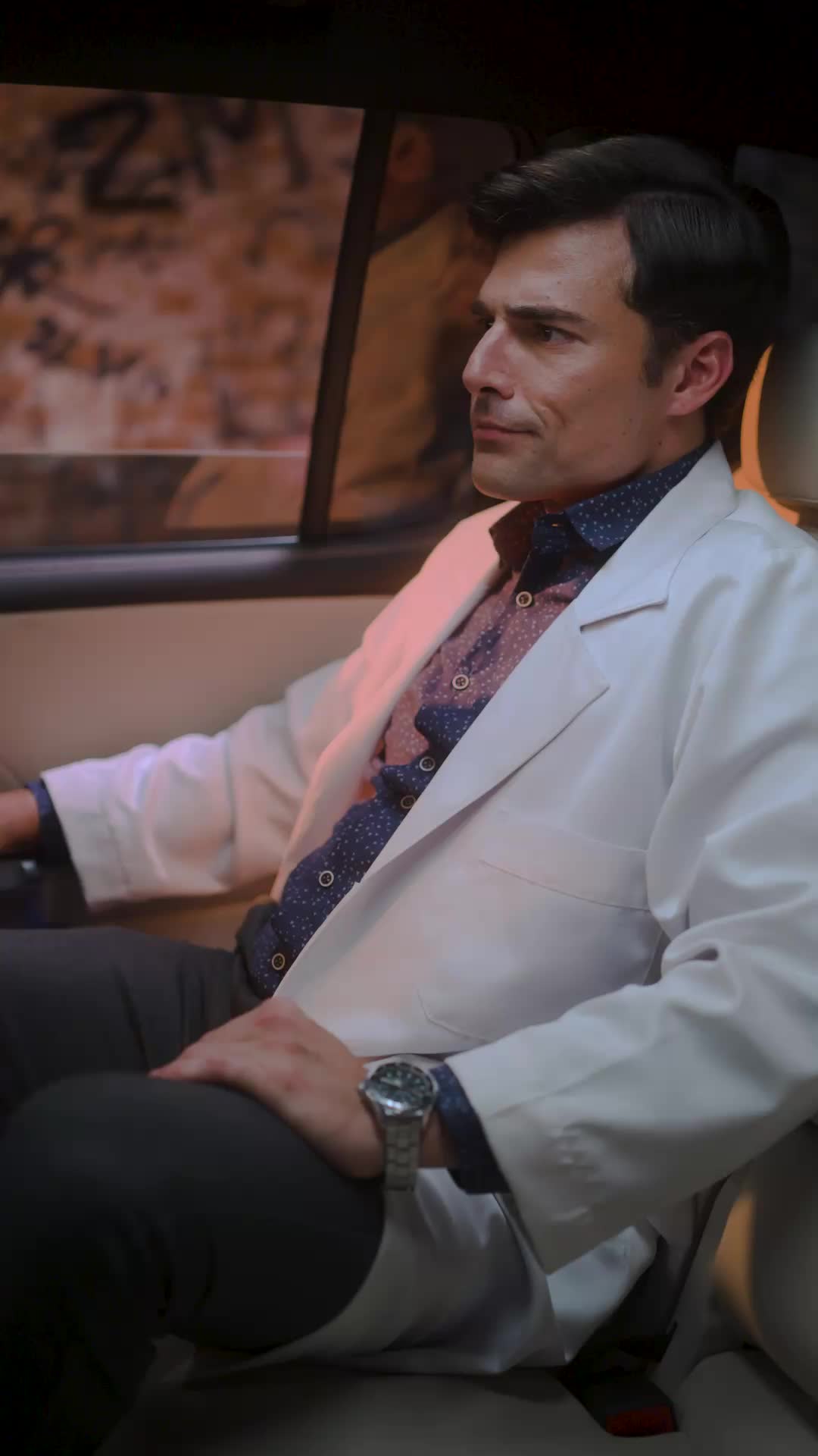 Léo sentado no banco de trás de um carro, de jaleco branco sobre camisa azul-escura estampada e relógio de aço, olhando pela janela para um muro pichado