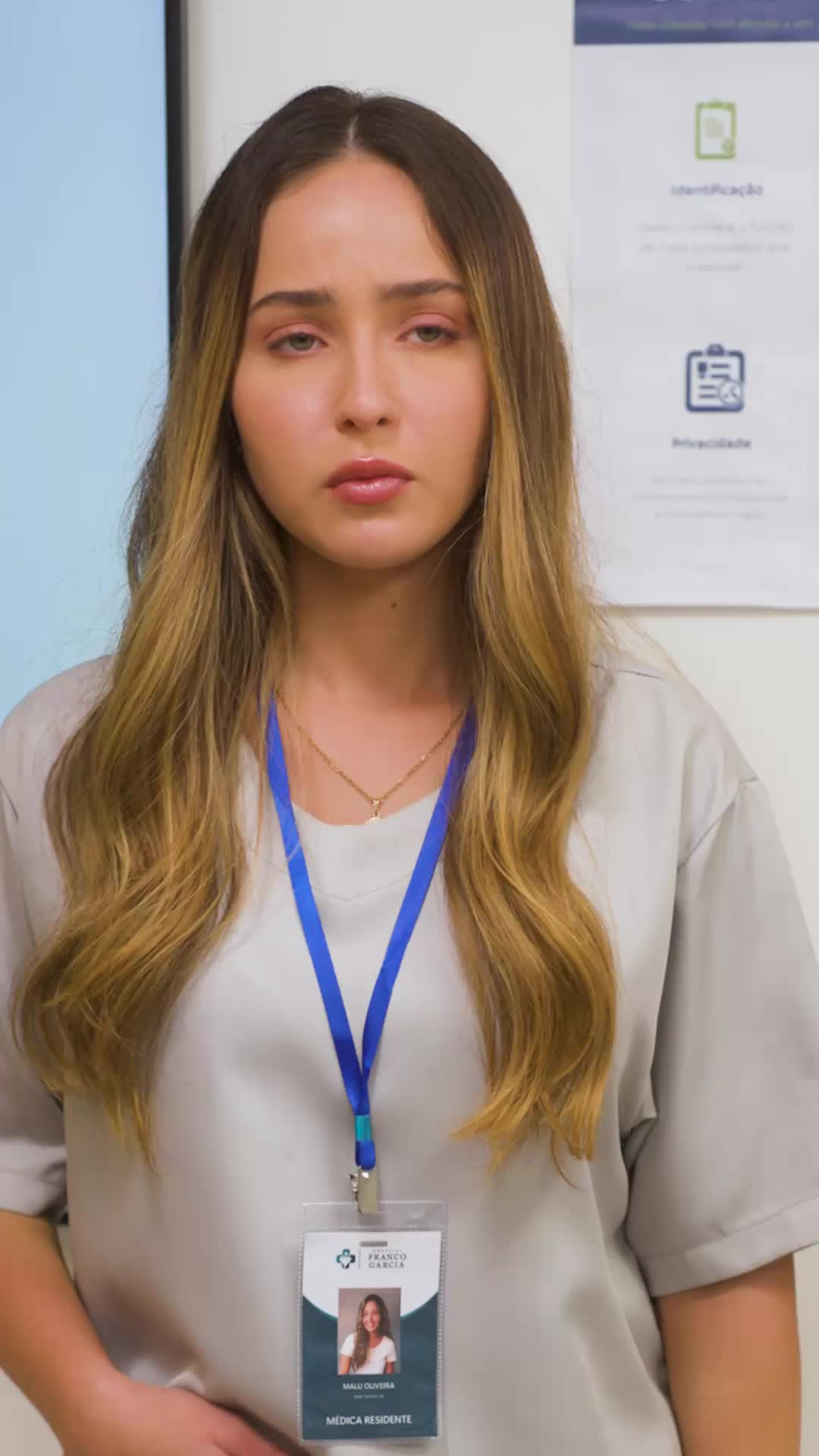 Malu de uniforme cinza, cabelo solto e corrente fina no pescoço, com o crachá de médica residente do hospital Franco Garcia, olhando para a frente com expressão séria