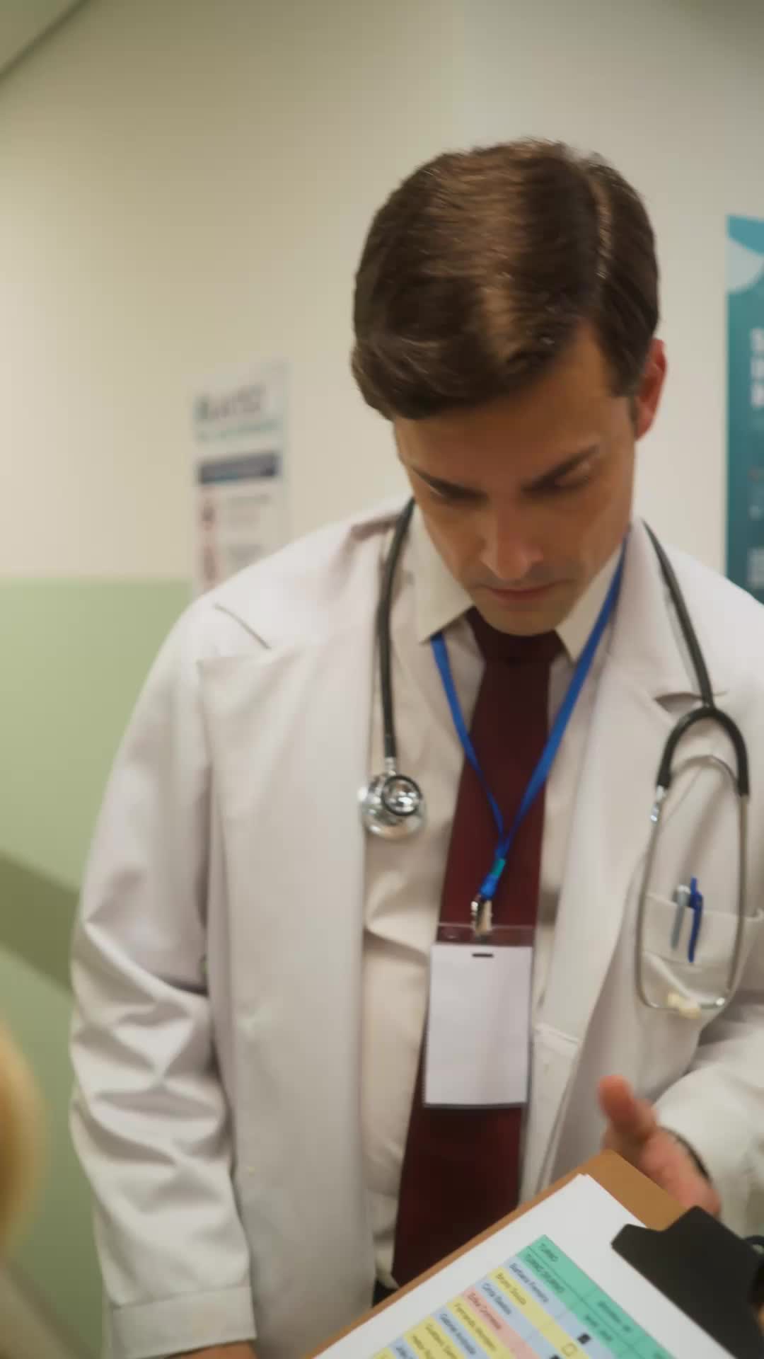 Léo de jaleco branco, gravata vinho e estetoscópio, com crachá em cordão azul, olhando para baixo para uma prancheta de prontuário num corredor de hospital de parede verde
